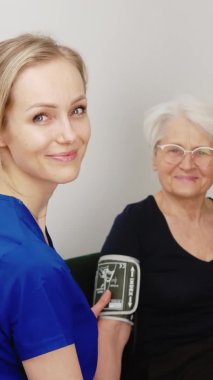 Mavi üniformalı neşeli genç hemşire ev ziyareti sırasında yaşlı gri saçlı kadının baskısını ölçüyor..