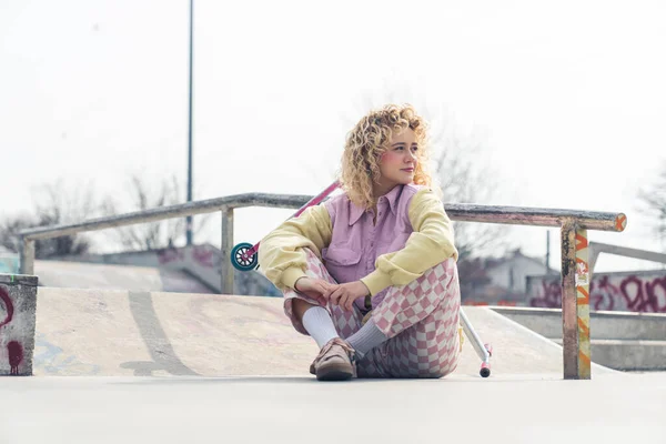 Ganska ung blond ruddy hipster kvinna sitter på skateboard marken leende, benen korsade, tittar bort rosa skoter på bakgrunden full skott kopiera utrymme — Stockfoto