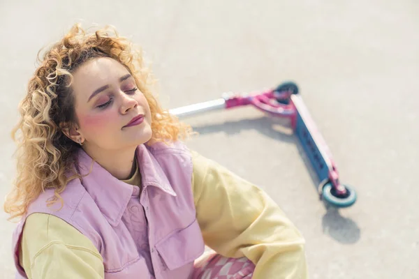 Muito jovem loira mulher rubicunda sentada no chão de esportes ao lado de uma scooter rosa ativo tempo livre conceito olhos fechado espaço cópia retrato — Fotografia de Stock