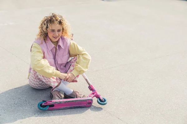 Muito jovem loiro encaracolado hipster mulher se senta no chão sorrindo, pernas cruzadas, scooter rosa na frente dela olhando afastado tiro completo cópia espaço cinza fundo — Fotografia de Stock