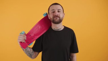 Orta yaşlı beyaz bir adam başının arkasında kırmızı bir kaykay tutuyor ve sarı arka planda kameraya bakıyor..