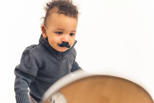 Adorable bébé garçon biracial drôle avec fausse moustache noire se regardant dans le miroir sur fond blanc. Plan studio. — Photo