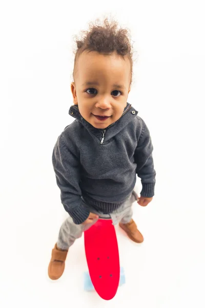 Top view of a curious playful biracial toddler boy standing over red skateboard wearing warm clothes. Vertical studio shot with white background. — Photo