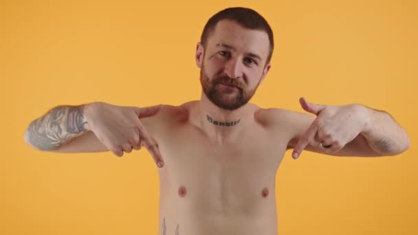 Barba topless tatuato ragazzo punta con entrambe le dita indice verso il basso, sorridente, e guardando la fotocamera su sfondo giallo. — Video Stock