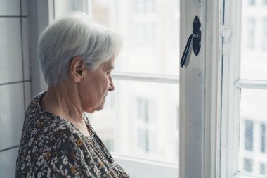 European elderly retired woman next to the window sadly gazing and thinking about her youth
