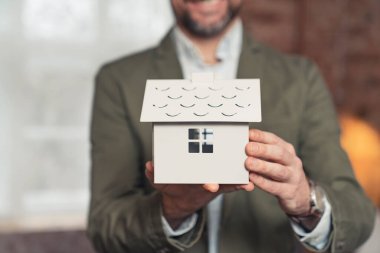 Takım elbiseli genç sakallı mimar elinde beyaz ev modeli bir gayrimenkul tutuyor.