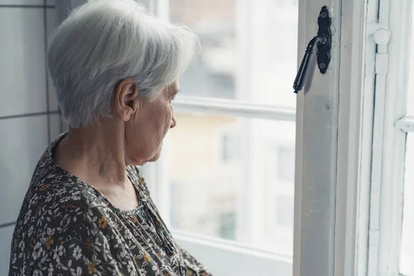 Miserable grey-haired older woman looking at the street throughout the window glass — Stockfoto