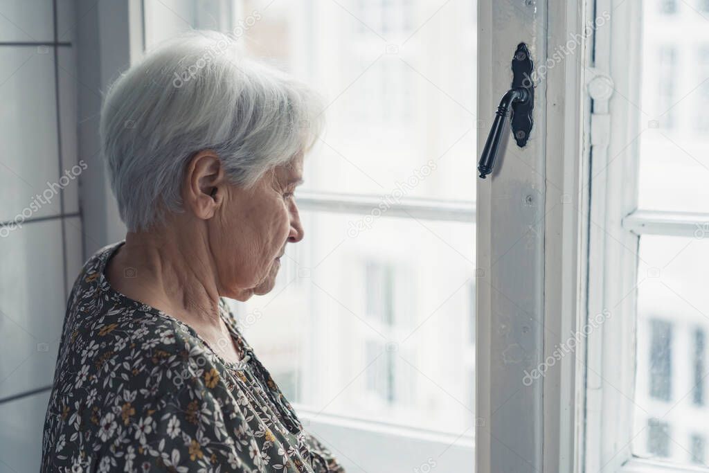 European elderly retired woman next to the window sadly gazing and thinking about her youth