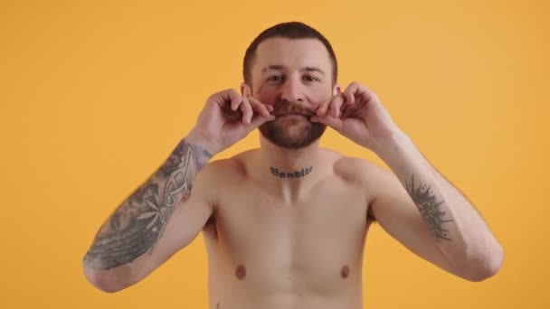 Shirtless Europeu tatuado cara em seus 30 anos tocando e brincando com seu bigode marrom arbusto enquanto olha para a câmera sobre fundo amarelo. — Vídeo de Stock