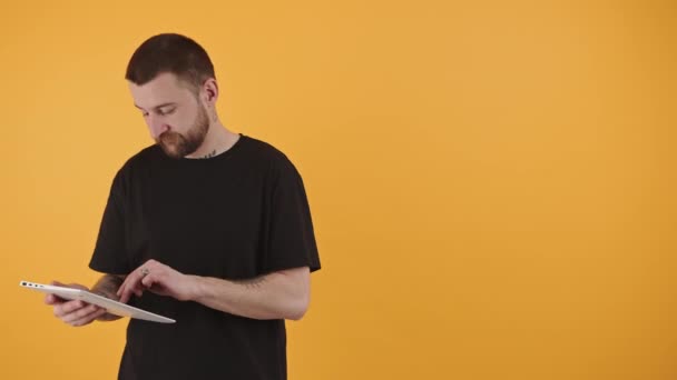 Kaukasische bärtige Macho tätowierte Kerl mit Tablet für Online-Shopping oder Unterhaltung über gelbem Hintergrund. Studioaufnahme. — Stockvideo