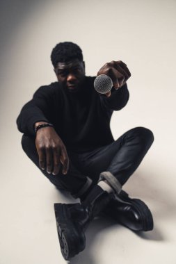 Beyaz arka planda oturan renkli adam mikrofonu kameraya doğrultuyor. Tam kare. 