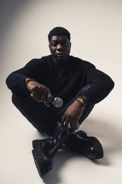 Beyaz arka planda oturan mikrofonlu siyah adam kameraya bakıyor. Tam kare.