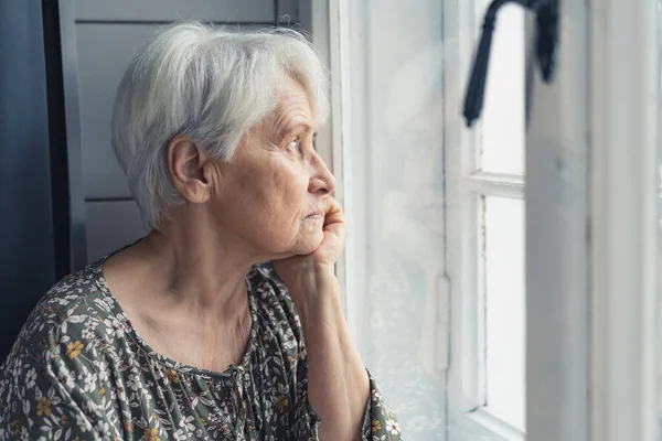 An elderly woman pensioner looks out the window sitting in his house — Stockfoto