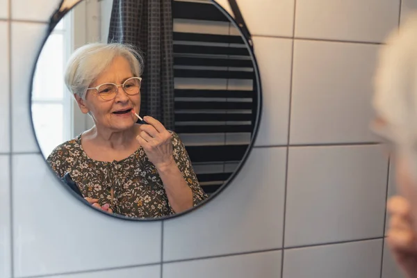 European grey-haired grandmother fixing her makeup in the mirror — стоковое фото