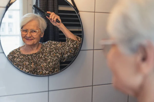 Пожилая седовласая женщина расчесывает волосы и смотрит на свое отражение в зеркале — стоковое фото