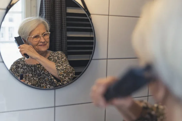 Elderly caucasian woman combs her hair and looks at the mirror — стоковое фото