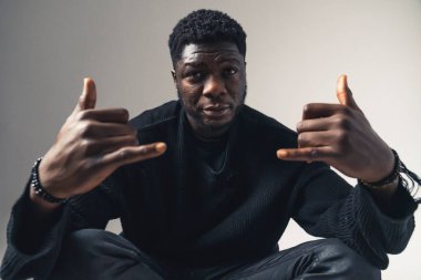 Shaka tabelasıyla oturmuş, beyaz arka plana bakıyor. Portre çekimi.