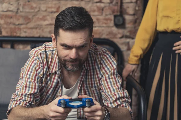 Kaukasische man in een unbuttoned gecontroleerd shirt het spelen van games met blauwe game controller en het negeren van zijn vrouw vriend — Stockfoto