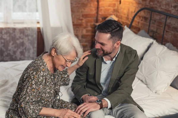 Kaukasiska gråhåriga pensionerad dam sitter på en säng med sin son och skrattar — Stockfoto