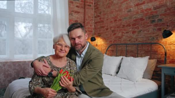 Anciana pensionista y su hijo milenario sentados en una cama, abrazando y celebrando el Día de las Madres — Vídeo de stock