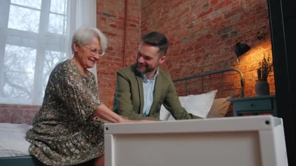 La abuela anciana europea y su hijo milenario balancean la cuna blanca donde un bebé pequeño está acostado — Vídeo de stock