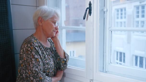 Cabelos grisalhos triste chateado mulher idosa em seus 60 anos olhando para fora da janela médio close-up interior tiro luz natural — Vídeo de Stock