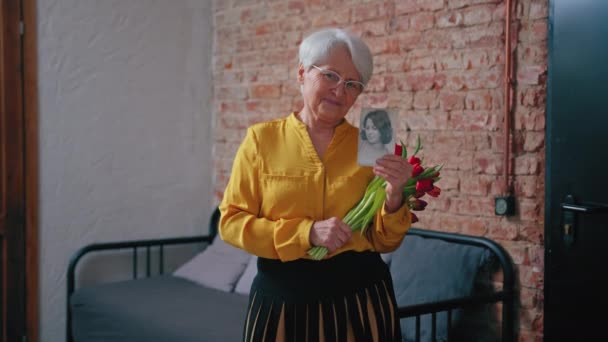 Usmívající se starší dáma s krátkými šedými vlasy porovnávající její současnou tvář s fotkou svého mladšího já — Stock video