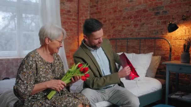 Ondernemer van middelbare leeftijd viert zijn moeders verjaardag en geeft haar een boeket rode tulpen en een geschenk — Stockvideo