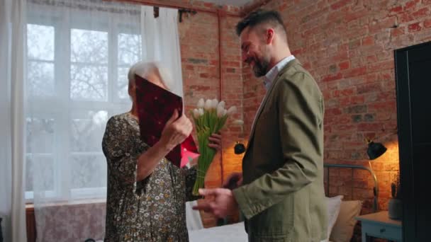 Dos personas caucásicas en un moderno piso celebrando el Día de las Madres, anciana recibiendo flores blancas y regalo de su hijo de mediana edad — Vídeos de Stock