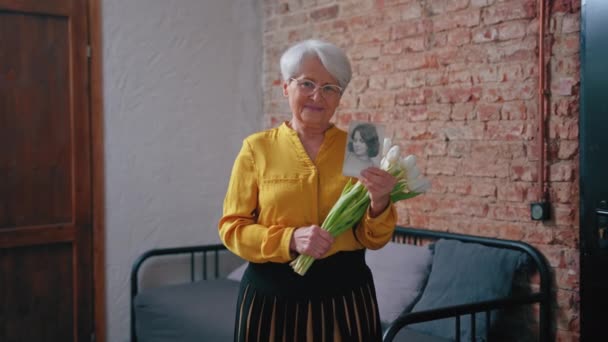 Kaukasische Rentnerin mit einem Strauß weißer Blumen und einem Schwarz-Weiß-Porträt aus ihrer Vergangenheit — Stockvideo
