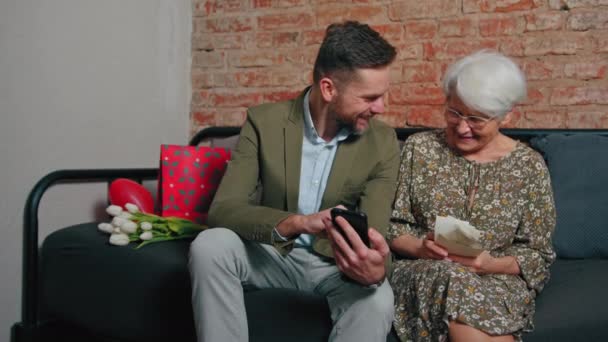 头发灰白的老年养恤金领取者妇女坐在沙发上，看着她千年大的儿子们的智能手机 — 图库视频影像