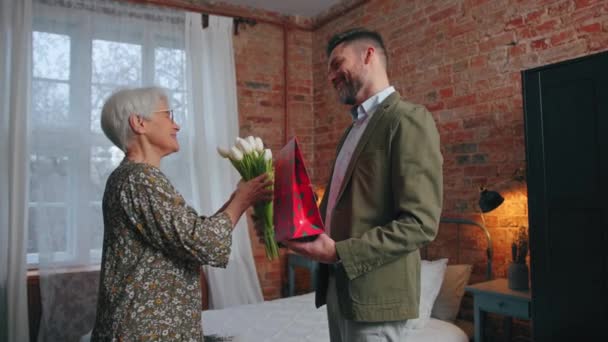 Joven empresario europeo llega a casa para sorprender a su anciana madre con ramo de flores y un regalo en el Día de las Madres — Vídeos de Stock