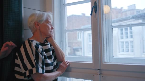 Cinza de cabelos caucasiano aposentado senhora olhando, infelizmente, para fora da janela médio close-up tiro interior — Vídeo de Stock