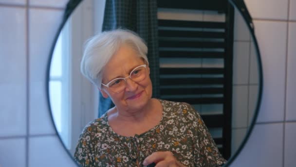 Пожилая седовласая женщина расчесывает волосы и смотрит на свое отражение в зеркале — стоковое видео