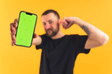 Sakallı, bıyıklı genç bir adam tek eliyle bir yumruk tutuyor ve ikinci el sarı arka plan stüdyosu ile cep telefonunu gösteriyor.