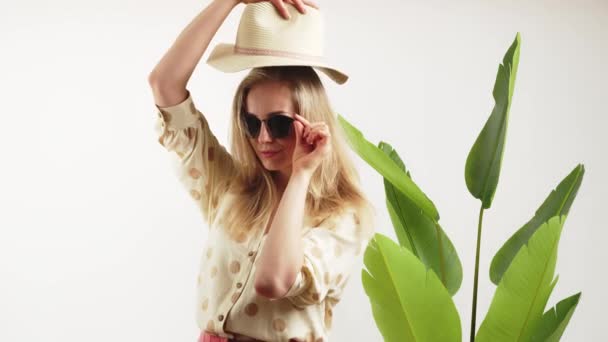 Vakre, hvite, lyshåret jente med svarte solbriller og hatt, som ser inn i kameraet hvit bakgrunn med grønne blader, tettsittende filmstudio – stockvideo