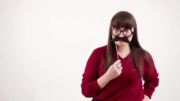 Νεαρή καυκάσια μελαχρινή να σιωπά κρατώντας ψεύτικο μουστάκι και γυαλιά μπροστά στο πρόσωπό της. — Αρχείο Βίντεο