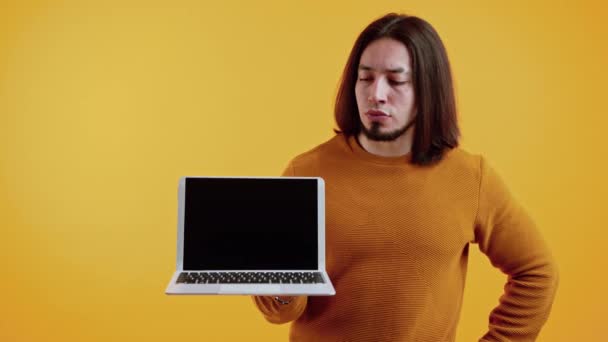 Enojado hombre caucásico barbudo con el pelo largo mirando a su ordenador portátil con pantalla negra simulada. Copiar espacio estudio filmado sobre fondo amarillo. — Vídeos de Stock