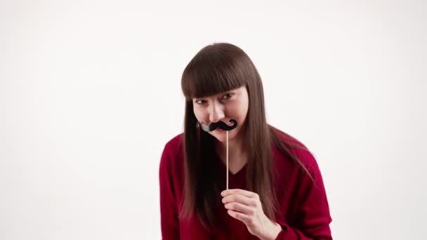 Забавна біла жінка дивиться на камеру і тримає підроблені вуса. Концепція Movember. Студійний знімок на білому тлі — стокове відео