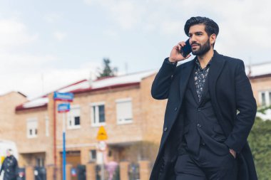 Siyah takım elbiseli şık Latin Girişimci ve kışlık ceketli akıllı telefondan yürüyor ve konuşuyor - düşük açılı çekim - banliyö bölgesi