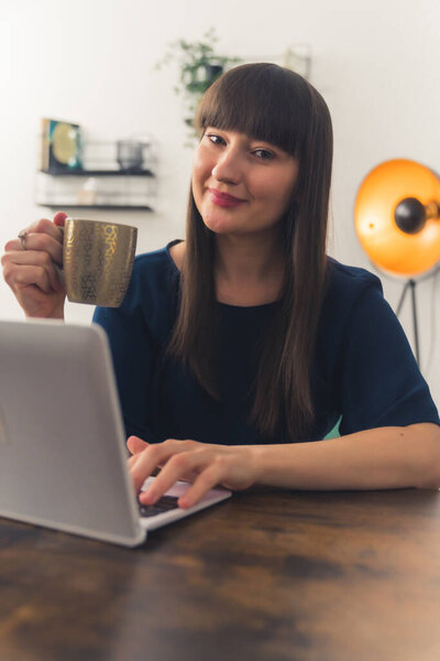 Pretty young Caucasian woman works at home, holding a cup of coffee, looking at the camera remote job concept copy space