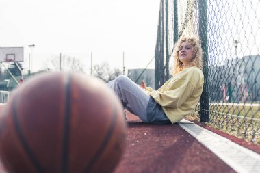  Çekici genç sarışın kadın basketbol sahasında oturuyor, yukarı bakıyor, basketbol topu önde sağlıklı yaşam tarzı konsepti fotokopi alanı. 