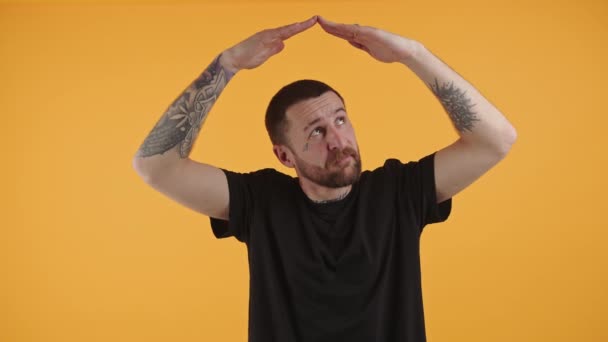 Handsome bearded man with tattoos making roof shape with hands above indoor studio shot orange on background medium shot — Stock video