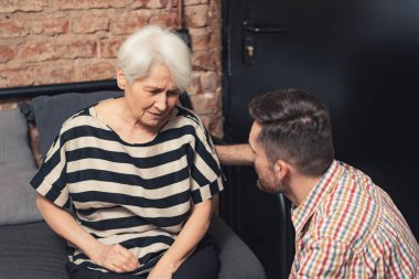 depressed elderly caucasian grandmother suffering from dementia sitting on a black couch and talking to her adult grandson