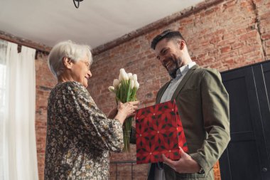 adult handsome son with facial hair giving red gift and white tulip bouquet to his elderly mother