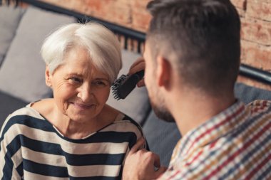 Mutlu emekli anne koltukta oturuyor ve saçlarını orta yaşlı oğlu tarıyor.
