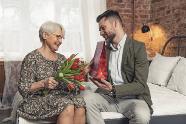 Yaşlı, kısa saçlı, zarif elbiseli anne orta yaşlı, beyaz iş adamı oğlundan bir hediye alıyor.