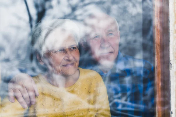 Senior elderly married couple embracing looking through the window during winter - love and togetherness at early age — Stockfoto