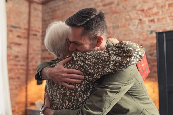 Kaukasiska tusenåriga entreprenör man gladeligen omfamna sin pensionerade gråhåriga mormor — Stockfoto