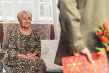 Orta yaşlı oğlu iyi dileklerini iletir ve yaşlı büyükannesi için bir hediye ve buket saklar.